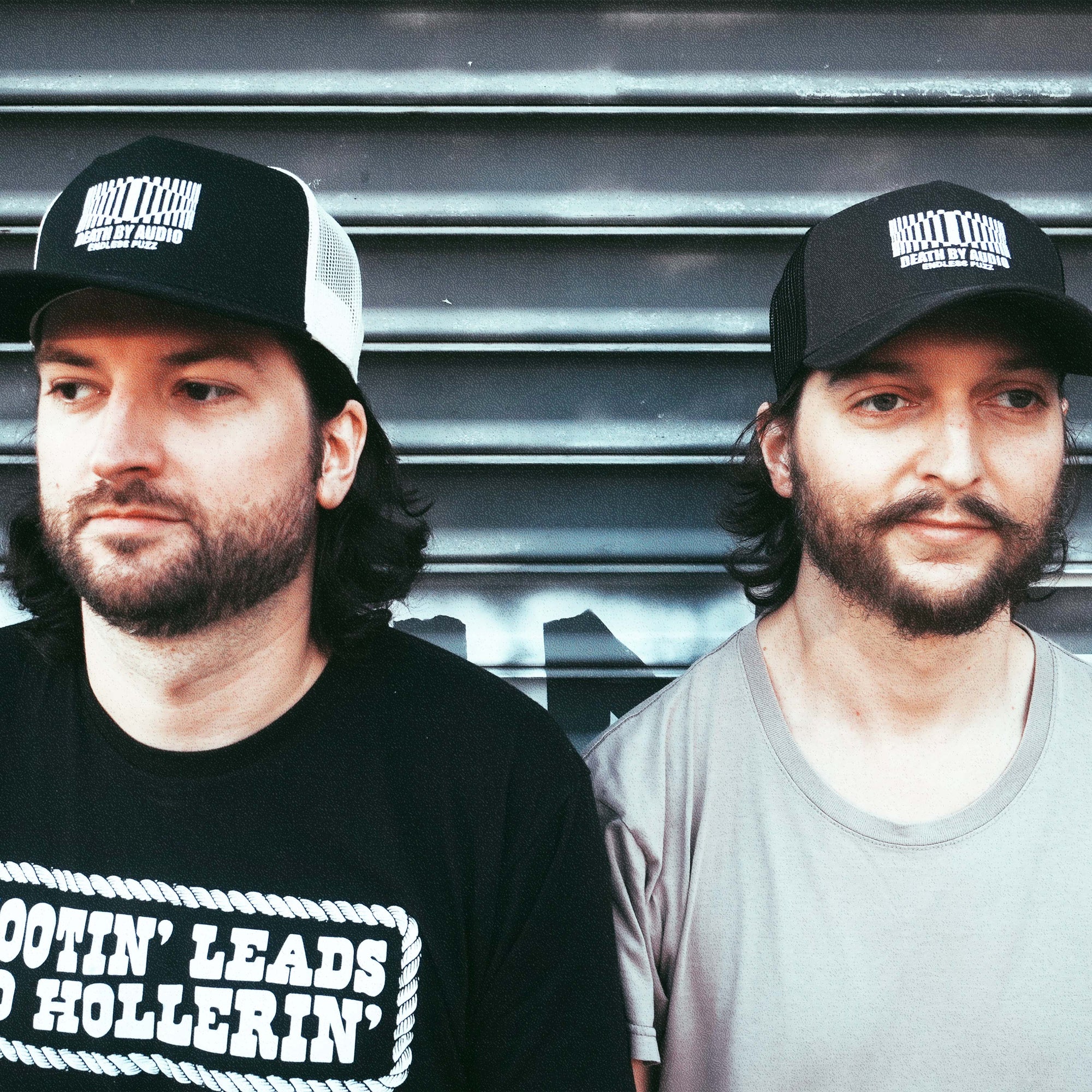 Leland and Cameron wearing the death by audio trucker hats in two color options. one is black with white mesh and the other is charcoal with black mesh.